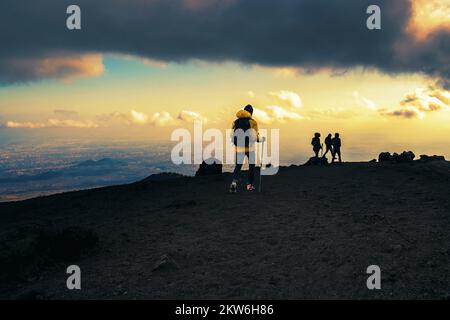 Gruppe von Wanderern, die durch den Lavastein des Ätna in Sizilien, Italien, bei Sonnenuntergang wandern - unbekannte Menschen, Blick von hinten in einem wolkigen Himmel - Stockfoto