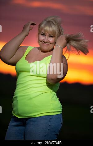 Eine dicke Frau mittleren Alters posiert in Jeans und einem hellen T-Shirt vor dem Hintergrund des Abendhimmels, übergewichtig xxl. Ein volles Mädchen genießt das Leben. Stockfoto