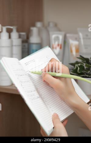 Die Hand einer Frau macht eine Notiz in ihrem Arbeitsbuch, auf einem Telefonschrank mit Kosmetik, einem Kosmetikbüro. Stockfoto