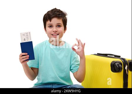 Ein niedlicher Junge im blauen T-Shirt, mit OK-Schild, sitzt neben dem gelben Koffer und hat eine Bordkarte in der Hand Stockfoto