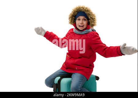 Ein fröhlicher Junge mit rotem Parka, ein süßes Lächeln vor der Kamera, sitzt auf einem blauen Koffer, reist am Wochenende ins Ausland Stockfoto