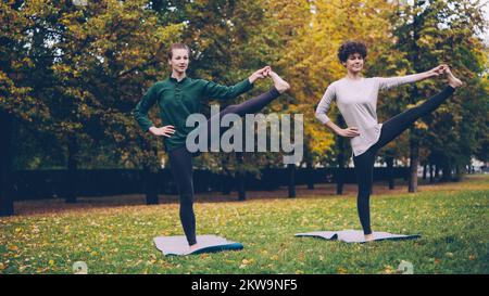 Schlanke junge Damen trainieren draußen im Park, stehen auf einem Bein und üben Padangusthasana während des individuellen Kurses mit erfahrenem Lehrer. Natur und Jugendkonzept. Stockfoto