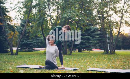 Die fleißige Yoga-Schülerin erlernt die Position des Kuhgesichts, sitzt auf der Matte und streckt Arme, während ihr ein erfahrener Lehrer hilft, Mädchen reden und lachen über Outdoor-Ptactice. Stockfoto