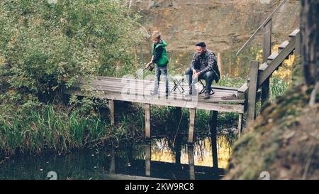 Gut aussehender Mann und sein süßer Sohn fischen im Teich vom Holzdock aus, sitzen auf Stühlen mit Stangen und reden. Liebende Familie, gemeinsames Hobby, Natur und Generationenkonzept. Stockfoto