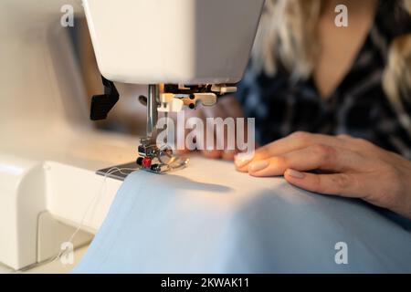 Junge Schneiderin näht Kleidung an Nähmaschinen. Nahtaufnahmen in der Werkstatt. Konzentrieren Sie sich auf Nähmaschinen und Taschentücher. Nähen eines kleinen Bu Stockfoto