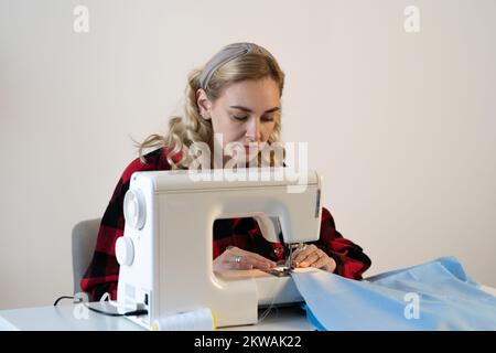 Fröhliche junge Frau näht, während sie an ihrem Arbeitsplatz in der Modewerkstatt sitzt. Die Näherin näht die Nähmaschine an. Schneiderei arbeitet an der Naht Stockfoto