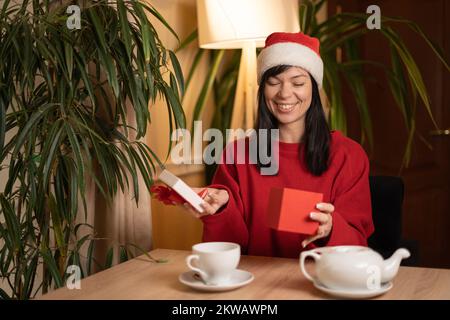 Junge, lachende brünette Frau mit weihnachtsmannmütze öffnet eine Schachtel mit einem Geschenk, lächelnd zufrieden mit einem guten Weihnachtsgeschenk. Speicherplatz kopieren Stockfoto