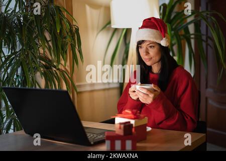 Wunderschöne junge Frau mit roter Mütze, die ihren Laptop benutzt, während sie im Wohnzimmer sitzt, Tee trinkt und sich online einen Weihnachtsfilm ansieht. Speicherplatz kopieren Stockfoto