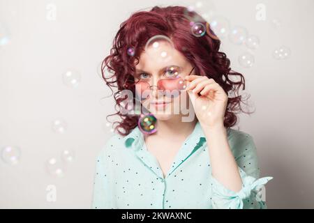 Eine Frau mit roten Locken, die eine herzförmige Sonnenbrille und Seifenblasen trägt Stockfoto