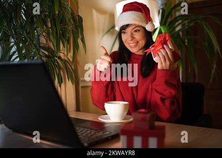 Weihnachtseinkäufe. Lächelnde glückliche Frau mit weihnachtsmannmütze, die eine Geschenkbox in der Hand hält und die Daumen nach oben zeigt. Frau mit Laptop sitzt zu Hause am Tisch mit Stockfoto