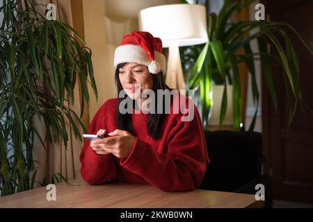 Positive Frau mit Weihnachtsmann-Hut sitzt am Tisch und benutzt das Handy im Haus an Heiligabend. Speicherplatz kopieren Stockfoto