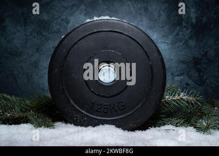 Hantelglockengewichtplatte und Weihnachtsbaumzweige auf Schnee. Fitness-Feiertage Winterzusammensetzung. Fitnessstudio Training Sporttraining Konzept. Stockfoto