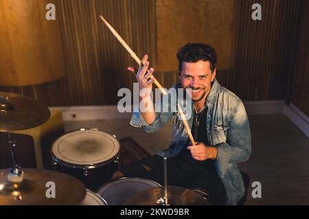 Ein weißer, bärtiger Drummer mittleren Alters hält Holzstäbchen, sitzt neben dem Schlag und lächelt vor der Kamera. Indoor-Aufnahme. Hochwertiges Foto Stockfoto