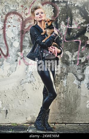 Er ist das perfekte Haustier für einen Rockstar. Das ganze Porträt einer gerissenen jungen Frau, die ihren kleinen Hund in einer urbanen Umgebung hält. Stockfoto