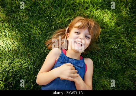 Spaß am sonnigen Tag. Ein süßes kleines Mädchen, das lächelt, während es sich auf dem Gras hinlegt. Stockfoto