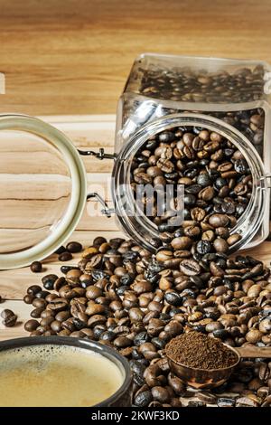 Eine Tasse frisch gebrühten Kaffees auf einem Holztisch gefüllt mit umgedrehten Kaffeebohnen aus einer Dose und einer Kupferuhr mit gemahlenem Kaffee Stockfoto