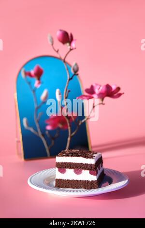 Schokoladenkuchen mit Sauerkirschen. Ein Stück Kuchen auf einem Teller mit Gabel. Süßes Dessert auf pinkfarbenem Hintergrund mit lebhaften Magnolienblüten. Stockfoto