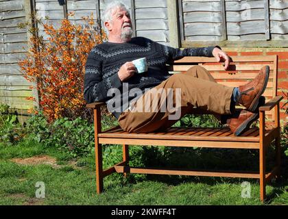 Mann sitzt mit erhobenen Füßen auf einer Bank und trinkt eine Tasse Tee oder Kaffee. Entspannende Pause. Stockfoto