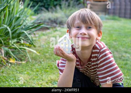 Kleines Haus in jungen Knabenhänden vor grünem Hintergrund von einem Baum in einem Garten. Stockfoto