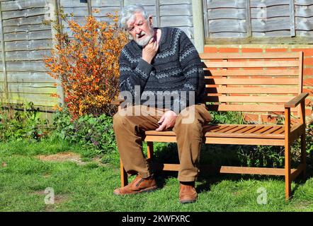 Senioren oder Senioren, die auf einer Bank sitzen. Nacken wegen Schmerzen halten. Möglicherweise Arthritis. Stockfoto