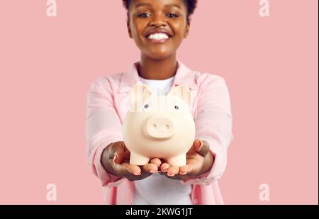 Portrait einer glücklichen, lächelnden afroamerikanischen Frau, die ein Sparschwein in den Händen hält, auf pinkfarbenem Hintergrund. Stockfoto