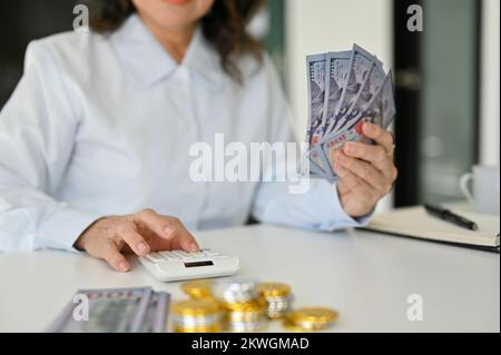 Zugeschnittenes Bild, eine asiatische Frau im Alter, die einen Taschenrechner benutzt, US-Dollar-Banknoten in der Hand hält und ihren finanziellen Anlagegewinn berechnet. Stockfoto