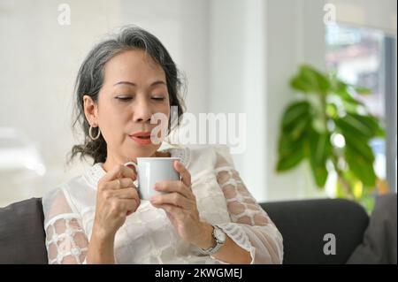 Eine friedliche und entspannte ältere asiatische Frau trinkt gerne morgens Kaffee, während sie in ihrem Wohnzimmer zu Hause chillt. Stockfoto