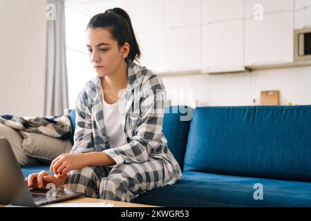 Eine junge Frau trägt einen Pyjama und verwendet einen Laptop, während sie zu Hause auf dem Sofa sitzt Stockfoto