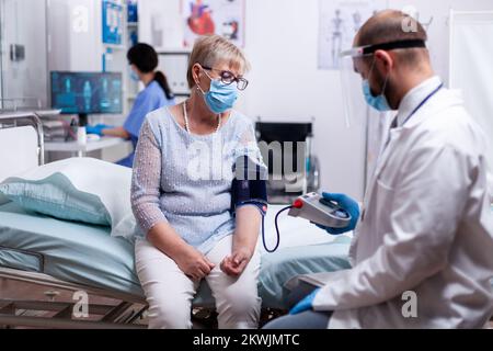 Arzt mit Gesichtsmaske gegen Coronavirus, der den Blutdruck der älteren Frau im Untersuchungsraum des Krankenhauses nimmt. Medizinische Kontrolle für Infektionen, Krankheit und Diagnose. Stockfoto