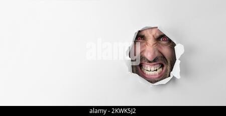 Wütender, verrückter Mann, der durch das Loch blickt, mit roten Augen auf weißem Hintergrund, mit Platz für Werbetexte Stockfoto