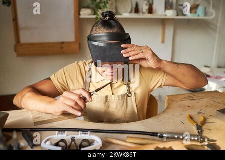 Vorderansicht einer Juwelierfrau, die eine Lupenbrille trägt und handgefertigte Kunstwerke in einem rustikalen Studio kreiert Stockfoto