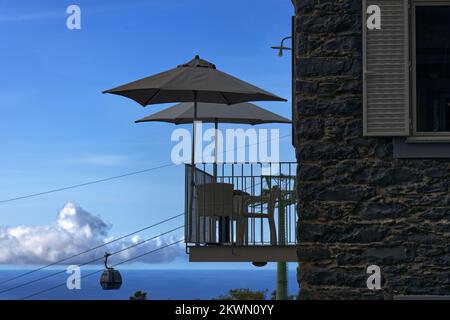 Funchal, Insel Madeira, Portugal - Sonnenschirme auf einem hohen Café-Balkon am Monte mit Seilbahn, die sich der Seilbahnstation im Hintergrund nähert Stockfoto