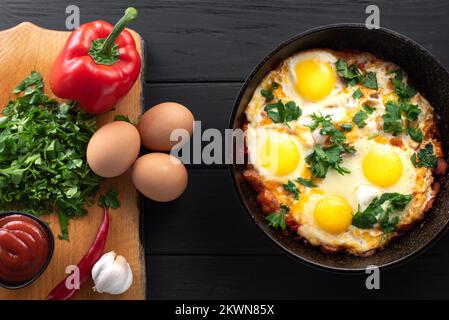 Shakshuka in einer Eisenpfanne mit Zutaten. Traditionelles Gericht aus dem Nahen Osten. Spiegeleier mit Tomaten, Paprika, Gemüse und Kräutern Stockfoto