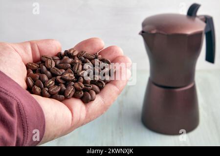 Frisch geröstete Kaffeebohnen auf einer gestreuten männlichen Handfläche vor dem Hintergrund einer braunen Geysirkaffeemaschine Stockfoto