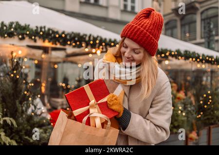 Stilvolle junge, wunderschöne, glückliche Frau geht mit Einkaufstaschen und Geschenkboxen auf der Straße der Stadt spazieren. Fröhliche attraktive Shoppingfrau mit Papiertaschen und Stockfoto