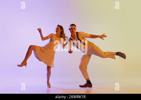 Ein hübsches Mädchen und ein Mann in Retro-Kostümen, die Brandtanz tanzen, isoliert auf fliederfarbenem Hintergrund in Neonlicht. Kunstbegriff Stockfoto