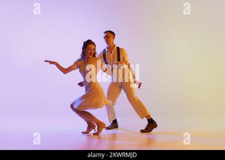 Brandtanz. Ein emotionales Paar Tänzer in Retro-Outfits tanzen soziale Tänze, isoliert auf einem abgestuften lilafarbenen Hintergrund in Neonlicht Stockfoto