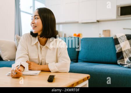 Junge süße lächelnde asiatische Frau, die zu Hause in einem gemütlichen, geräumigen Wohnzimmer Notizen schreibt Stockfoto