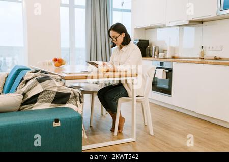 Junge süße lächelnde asiatische Frau in Brille mit Tablet, die zu Hause am Tisch in der Küche arbeitet Stockfoto