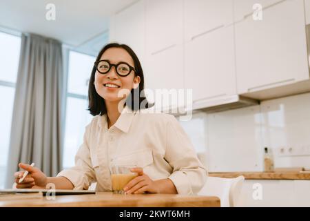 Junge süße lächelnde asiatische Frau mit Brille, die zu Hause am Tisch in der Küche arbeitet Stockfoto