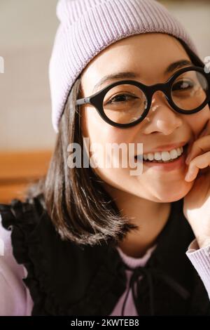 Junge Asiatin mit Brille, die lächelt und drinnen in die Kamera schaut Stockfoto