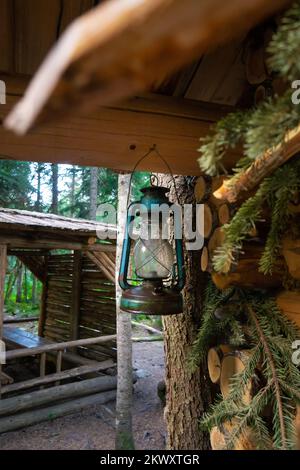 Alte Kerosin-Laterne, die an der Ecke eines Holzblockhauses hängt und neben einem Pavillon im Wald steht. Vertikales Foto Stockfoto