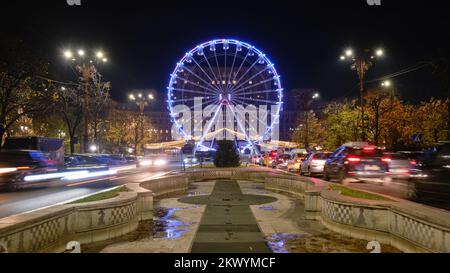 Bukarest Riesenrad für den Weihnachtsmarkt, vor dem Parlamentsgebäude, mit Autos, die zur Hauptverkehrszeit vorbeifahren. Nacht, Licht, beleuchtet, symmetrisch Stockfoto