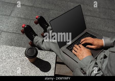 Draufsicht auf einen verkürzten Geschäftsmann in Rollschuhen, der auf einem Laptop tippt, mit leerem Bildschirm in der Nähe von Kaffee, um ins Freie zu gehen, Stockbild Stockfoto