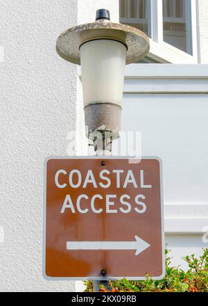 Vertikales Schild für den Küstenzugang an einer Laterne in La Jolla, Kalifornien. Nahaufnahme einer Beschilderung vor dem weißen Gebäude mit Geländern und Sträuchern draußen. Stockfoto