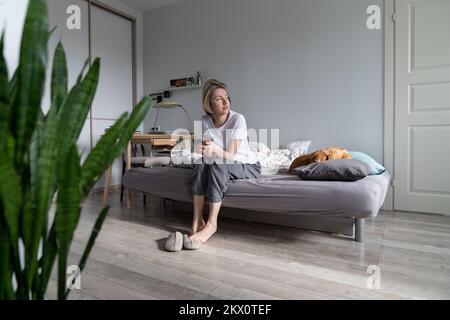 Schläfrige Frau, die auf dem Bett sitzt und ein Handy hält, den Wecker ausschaltet und mit einem Hund aufwacht Stockfoto