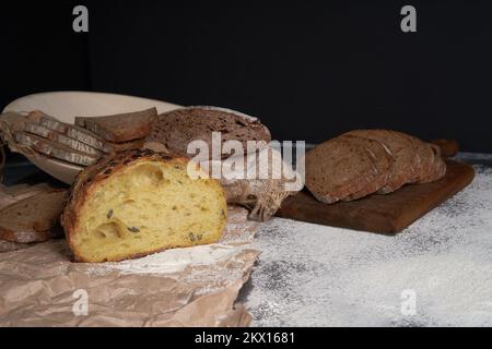 Verschiedene Brotsorten und ein Laib auf Holzhintergrund. Verschüttetes Mehl. Vorderansicht. Speicherplatz kopieren. Ein halbgeschnittener Laib. Auf schwarzem isoliertem Hintergrund. Frisches, verkrustetes Brot. Stockfoto