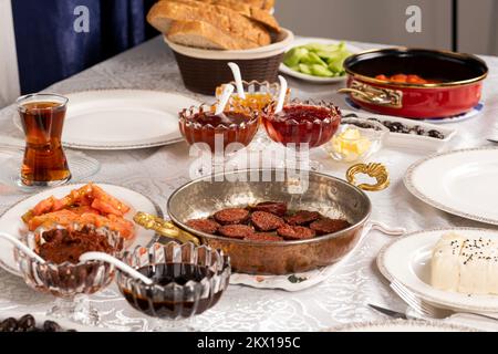 Reichhaltiger Frühstückstisch. Türkische Wurst namens Sucuk in Bratpfanne, fertig zum Essen. Brot, Käse, Tomaten, Marmelade, Honig, Tee im Glas. Stockfoto