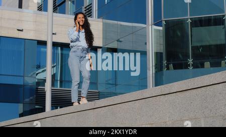 Junge, fröhliche, schöne, brünette Geschäftsfrau, Gewinner eines sorgenfreien Mädchens, das auf einem urbanen blauen Glasgebäude im Hintergrund läuft und mit dem Handy tanzt Stockfoto