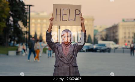 Trauriges, einsames, armes muslimisches Mädchen trägt Hijab Frau steht in der Stadt und schaut auf die Kamera zeigt zeigt zeigt, dass Pappbanner Text Hilfe braucht Schutz Arbeit Stockfoto